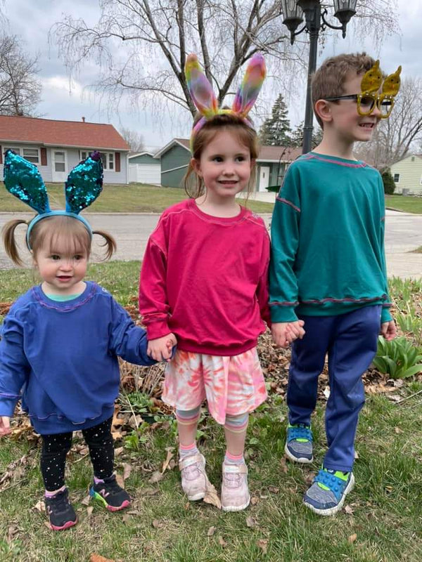 Three children wearing colorful outfits and headbands standing outdoors on a residential street.